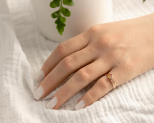 Hand wearing 14K yellow gold birthstone stackable rings with 2mm round stones sizes 4-10 on a white fabric with green plant in background  
